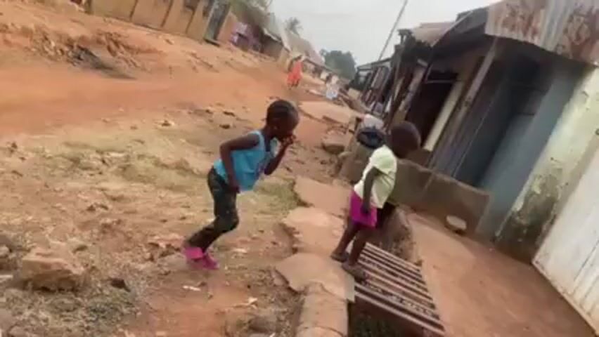 Two young children are playing in a dirt road in a village or town. The older child, wearing a blue shirt and camo leggings, is standing on a small concrete ledge next to a drainage ditch with a metal grate. The younger child, wearing a yellow shirt and pink shorts, is running across the grate. A chicken runs out from under the grate. The setting is a narrow dirt street with simple buildings lining the sides. A few people can be seen walking in the background.