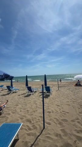 A full shot shows an empty beach on a bright sunny day, with a couple of beach loungers and two closed blue beach umbrellas in the left foreground. In the mid-ground right, a white beach umbrella is open, with a person sitting under it facing the ocean. A life preserver attached to a blue pole is also in the mid-ground right. The ocean is bright blue with white waves coming to shore. The sand is light brown. The sky is bright blue with some white clouds. The camera zooms in on the ocean.