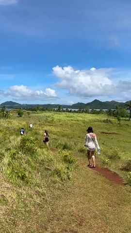 The video shows a group of people walking along a dirt path through a grassy field on a sunny day.  The field is surrounded by trees and hills in the distance, and the sky is blue with scattered white clouds.  One person is walking in the foreground, wearing a white dress and a black hat. Three other people are ahead on the path, two wearing shorts and one wearing a blue shirt.