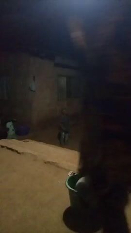 A dimly lit outdoor scene reveals a person in a patterned dress walking away from the camera towards a slightly elevated area where several other individuals are present. The surroundings appear to be a village setting at night, with modest structures visible in the background. A green bucket sits in the foreground. The people in the background are moving about, though their specific actions are unclear due to the low light and distance from the camera.