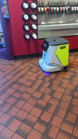 A small, white and yellow robot with a screen displaying two zeros is maneuvering on a tiled floor. The robot has a black top with a red button. To the left is a purple vending machine and to the right is a red wall with cup dispensers.