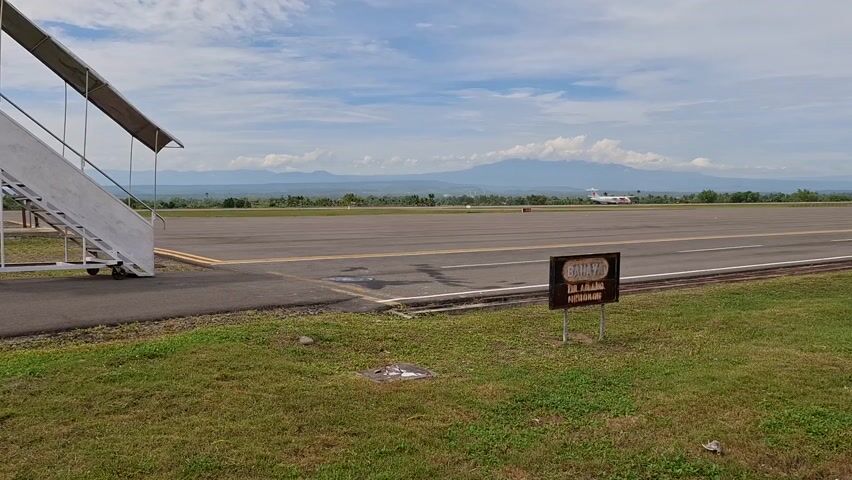 The video presents a static, wide shot of an airport runway area on a bright, partly cloudy day. In the foreground on the left, a set of white portable aircraft stairs stands on the paved edge of the tarmac, next to a patch of green grass. To the right, a small brown sign with black text is planted in the grass. The sign, written in Indonesian, clearly reads "BAHAYA" (Danger) at the top, followed by "DILARANG MELINTAS" (Forbidden to cross), and "LANDASAN PACU" (Runway), serving as a warning.

The main runway stretches across the middle of the frame, a wide expanse of asphalt with white and yellow markings. In the far distance on the runway, a white airplane can be seen, appearing to be stationary or slowly taxiing. Beyond the runway, a landscape of green trees and fields extends to the horizon, where several large mountains are visible under the partially clouded sky. The overall scene suggests a regional airport with natural surroundings, likely in Panigah, Indonesia, as indicated by the context. The time of day appears to be midday or early afternoon, characterized by clear visibility and ample sunlight.