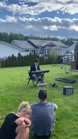 A young man in a black jacket and white shirt plays a keyboard set up on a chair in a grassy yard. In front of him, two people sit on the grass, facing away from the camera, watching him play. A small amplifier sits to the right of the keyboard. Behind the performer, a row of houses with modern architecture and dark roofs are visible, bordered by a line of evergreen trees. A greenhouse and a trailer are also present in the background. The sky is partly cloudy with large, dramatic white clouds against a blue sky, suggesting late afternoon or early evening. The overall mood appears relaxed and pleasant, with the audience enjoying a private outdoor performance. The location context indicates this scene takes place in Joensuu, Finland.