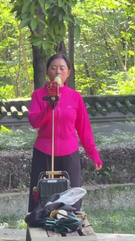 A woman is captured singing into a green microphone in an outdoor setting, likely a park or garden. She is standing, dressed in a bright pink long-sleeved athletic jacket with a white logo on the right chest, and dark, wide-legged pants. Her expression is focused, suggesting engagement with her performance.

The microphone is connected to a golden-colored stand that also holds a red smartphone. The phone's screen is visible, displaying Chinese characters, likely lyrics or a karaoke application interface. Below the phone stand, on a wooden surface, sits a black portable speaker or amplifier unit. Alongside it, sunglasses and other personal items are placed.

The background is rich with greenery, featuring trees and dense bushes, indicating a natural environment. Behind the foliage, a traditional Chinese-style wall with dark, curved roof tiles can be seen, adding a cultural element to the scene. The lighting suggests it is daytime, possibly bright and sunny. The setting and activity point to an outdoor karaoke or singing performance in a public space in Banan, China.
