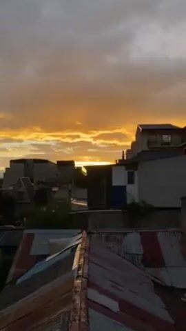 The video presents a dynamic outdoor shot of a cityscape during what appears to be sunset or sunrise. The sky is filled with orange-yellow light breaking through clouds. In the foreground, various building rooftops are visible. They feature different materials like corrugated iron. Behind these rooftops, there are multi-story buildings of varying architectural styles, some with visible windows. A few trees and urban infrastructure elements such as power lines can also be seen. The camera pans across the scene, providing a wider view of the surrounding cityscape and distant mountains.