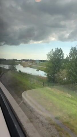 The video shows a view from a moving train in daylight. Outside the train window is a canal, lush green grass, trees, and a building in the distance. A road runs parallel to the canal. The sky is mostly covered by heavy, dark clouds, with some patches of blue visible in the distance. The train track runs close to the canal.  A railing is visible on the train track between the track and the canal.