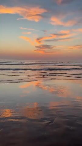 A vertical video captures a sunset over the ocean. The sky is filled with wispy clouds colored in shades of pink, orange, and blue. The ocean has gentle waves that create ripples on the wet sand of the beach in the foreground. The wet sand perfectly reflects the colors of the sky and the clouds, creating a mirror-like effect. The scene is peaceful and serene.