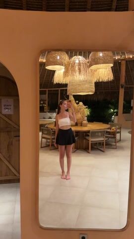 A young woman is taking a selfie in front of a large mirror in what appears to be the lobby of a resort. She is fair-skinned with long brown hair, wearing a white crop top and a short black skirt. She's holding a smartphone, obscuring her face. The lobby has a high, thatched roof and is lit by large, woven pendant lights. There is a large round wooden table with chairs in the center of the room, and other seating areas are visible in the background. The floor is light-colored tile. A door with a "STAFF ONLY" sign is visible to the left of the mirror. The overall atmosphere appears calm and luxurious.