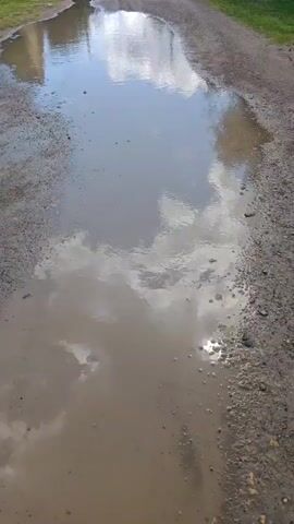 This video shows a very large, muddy puddle on an unpaved road. The puddle fills most of the road and reflects the sky, which has white clouds and blue patches. The road is lined with gravel and dirt. There are some indications of buildings or structures in the distance.
