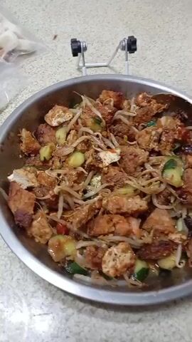 A close-up shot focuses on a dish served in a metal bowl on a light-colored countertop. The dish appears to be a mix of ingredients coated in a reddish-brown sauce. Visible components include chunks of what looks like fried tofu or possibly meat, bean sprouts, and sliced cucumber. A spoon is seen partially submerged in the dish in some frames. In the background, there's a white metal stand with black knobs and a plastic bag containing white items. The camera slightly pans across the food during the short clip. No people are visible, and no text is present in the video.