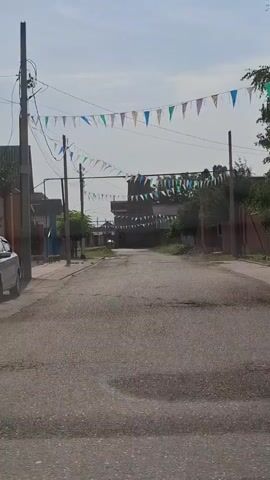 The video shows a street in a village or town. There are strings of colorful triangular flags strung across the street, attached to utility poles. The street is paved and has some potholes. On either side of the street are houses and fences. In the distance, there is a building with a tower, possibly a church or a mosque. The sky is partly cloudy and the overall impression is that of a sunny day. There are no people visible in the video.