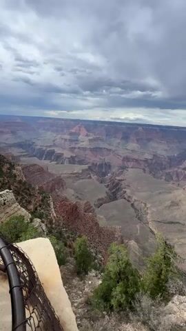 This is a video of a drone shot overlooking the Grand Canyon. The drone is flying over the edge of the canyon, looking out at the vast expanse of red rock formations. The sky is cloudy and overcast, but the sun is peeking through in some areas, creating a dramatic effect. The drone is moving slowly and steadily, giving the viewer a good sense of the scale and grandeur of the canyon. There are no people or animals visible in the video. The environment is rugged and natural, with sparse vegetation on the canyon walls. The video is filmed from a high vantage point, providing a breathtaking view of the geological wonder.
