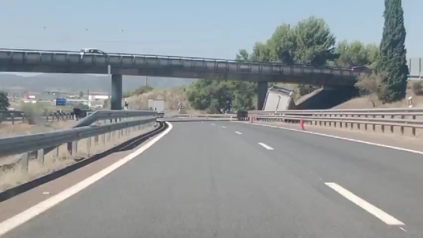 The video shows a view from a vehicle traveling on a highway. In the foreground, the highway is paved with asphalt and has white lane markings. A metal guardrail runs along the right side of the road. In the mid-ground, an overpass spans the highway. To the right of the highway, there is a tilted trailer of a semi-truck, indicating an accident. Orange traffic cones are visible near the trailer. A sign with a warning symbol and a silhouette of a tree is posted on the right side of the highway. In the background, there are hills covered with vegetation and some buildings. The sky is clear and blue. Several other vehicles, including trucks and cars, are visible on the highway and on the overpass.