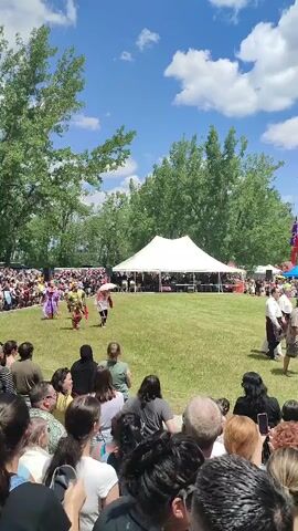 In this video, a group of people are gathered outdoors on a sunny day. The scene appears to be a cultural event, possibly a powwow or festival, with many spectators watching a performance. In the foreground, the backs of several audience members are visible. In the middle ground, a grassy area is set up for a performance. A white tent is visible in the background, likely serving as a stage or shelter. Several performers in traditional attire are walking across the grass, with some onlookers also visible in the distance. The overall environment is outdoors with trees and a blue sky with white clouds.
