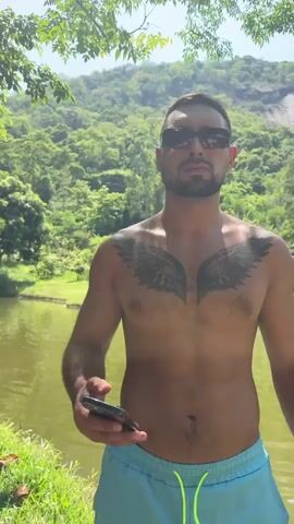 A man wearing sunglasses and blue swim trunks stands shirtless in front of a body of water. He has a tattoo of wings on his chest. He holds a smartphone in his left hand. The background is a lush green hillside with trees. A fountain is visible in the water behind him.