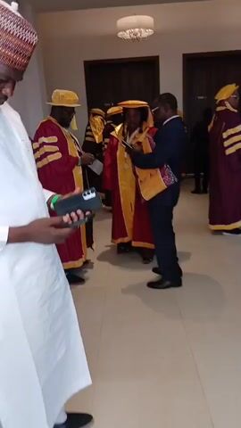 The video captures an indoor scene in Unguwan Sanusi, Nigeria, likely a hall or lobby with light-colored tiled floors, white walls, and dark wooden doors in the background. Two modern, circular ceiling light fixtures illuminate the space.

The primary subjects are a group of adults, most of whom are dressed in academic regalia. Their attire consists of deep maroon or purple robes with prominent yellow or gold sashes and trim, some featuring large white or cream-colored stripes on the sleeves. Many are also wearing matching academic hats, some resembling soft caps and others mortarboard style. One man is observed wearing a dark blue suit, interacting with individuals in robes.

The activity suggests a gathering related to an academic event, such as a graduation ceremony or convocation. People are seen standing, conversing, and several individuals are actively using their smartphones, looking at the screens or holding them up. One woman in regalia is wearing sunglasses and carrying a black handbag. The overall atmosphere appears calm and social as participants wait or mingle. There is no visible text in the scene. The lighting indicates it is daytime.