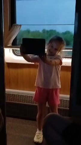 A young girl stands inside a train car, holding a tablet computer in front of her face and appearing to take a selfie. She is wearing a white t-shirt and pink shorts, with white clogs on her feet. The train car has wooden paneling on the walls and a window to her left, through which trees and a cloudy sky are visible. The girl's hair is pulled back in braids. The camera is focused on the girl and the tablet.