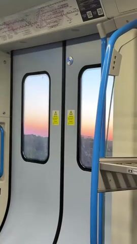 The video captures the interior of a moving train, focusing on the closed passenger doors and the view outside. The doors are light grey with black rubber seals, each featuring a small, rounded rectangular window. A silver circular button or handle is located where the doors meet. On each door panel, two yellow triangular warning stickers with black exclamation marks are visible, displaying text that includes "CAUTION."

Through the windows, a vibrant sunset or sunrise paints the sky with rich orange and pink hues near the horizon, transitioning to lighter blues higher up. Dark, silhouetted tree lines or distant structures are visible at the bottom of the windows. The continuous change in the outside scenery indicates the train is in motion.

Inside the carriage, prominent blue grab handles or poles are visible on the right, and a smaller blue handle is on the left. Above the doors, an overhead panel displays a detailed route map, characteristic of a regional or urban rail network, with various colored lines and station names. The train's interior lighting is soft, contrasting with the bright, warm glow of the sky outside. No people are visible in the shot. The scene evokes a tranquil moment of travel during either dawn or dusk.