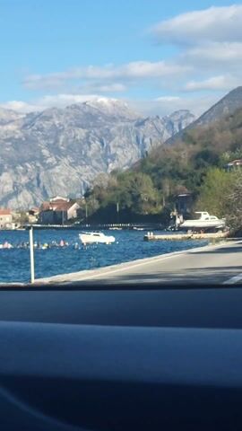 The video depicts a scenic view from inside a moving vehicle, looking out over a body of water toward a mountainous landscape. The water contains several boats, one of which is white and anchored. Buoys, appearing to be orange, yellow, and black, float on the water near the boats.  On the shoreline, there are a few buildings with tiled roofs and a small dock with another boat. The mountains in the background are grey and rocky with some snow visible on the peaks. Along the roadside, there's a sparse arrangement of trees with budding green leaves. The camera angle is low and focused through the car's windshield, with the dashboard visible at the bottom. The overall impression is one of a bright, clear day at a coastal location.