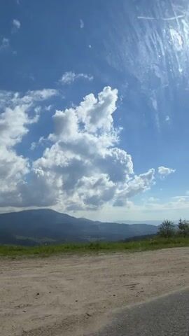 The video shows a view from a moving vehicle. The camera pans across a landscape of rolling hills and mountains under a blue sky with scattered clouds. Trees dot the green hillsides. The sun is visible in the upper right of the frame. The camera then moves to show the view from a window with a gray curtain.