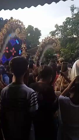 The scene shows a crowd of people gathered in an outdoor setting. Many are holding up their phones, seemingly recording or taking pictures of a large stage or structure adorned with golden decorations, lights, and possibly flags. Trees are visible in the background and the sky is overcast. It appears to be an event or celebration of some kind with a stage and lighting setup.