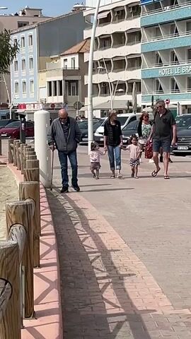On a sunny day, an elderly man using a cane and an older woman are walking on a sidewalk while each holding the hand of a young girl. The man is wearing a grey jacket and blue jeans, and the woman is wearing a black and white striped top and blue jeans. The girls are wearing simple tops and shorts. Another couple is walking behind them. The sidewalk is made of brick and borders a sandy area enclosed by wooden posts. Behind the group, a row of parked cars lines the street in front of buildings, one of which is labeled "HOTEL LE BR." The sky is clear and blue.