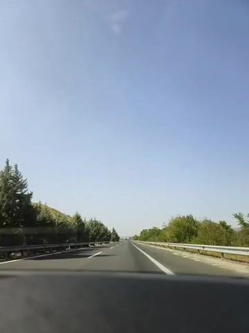 The video shows a POV shot from inside a vehicle traveling on a highway. The road is paved and has white lines marking the lanes. On either side of the road, there are guardrails and vegetation, including trees and bushes. The sky is clear and blue. The camera is positioned looking forward through the windshield. The video is short and shows the vehicle continuing along the highway without any significant events.