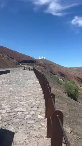 The video shows a view from a stone pathway with a metal railing on the right. The pathway leads up a hill towards a landscape dominated by volcanic terrain. In the distance, on the crest of a hill, are several white observatory domes. The ground is a mix of dark soil, reddish-brown volcanic rock, and sparse vegetation. A large, cone-shaped volcano with a plume of smoke or clouds near its summit is visible on the far right horizon. The sky is a clear, vibrant blue with a few wispy white clouds. The sun is shining, casting shadows.
