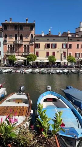 This media file captures a serene waterfront scene in Desenzano del Garda, Italy. The foreground is dominated by several small boats docked on the tranquil water. Two boats, one with a blue hull and another with a wooden deck, are prominently featured, with lush green plants and a few pink flowers adding a touch of natural beauty. Beyond the boats, a canal lined with colorful buildings stretches into the distance.

The buildings exhibit classic Italian architecture with pastel facades in shades of cream, salmon, and light blue, adorned with dark shutters and balconies. The architecture suggests a charming, historic European town. Along the waterfront, a row of restaurants and cafes is visible, with tables and chairs set up under umbrellas, indicating a leisurely atmosphere. People are seated at some of these tables, enjoying the pleasant weather and ambiance.

The time of day appears to be midday, with bright sunshine casting sharp shadows and a clear blue sky overhead, suggesting fair weather. The overall mood is peaceful and inviting, conveying a sense of relaxation and vacation. The presence of numerous small boats hints at a popular spot for leisure boating or as a picturesque harbor. No visible text is present in the media file.