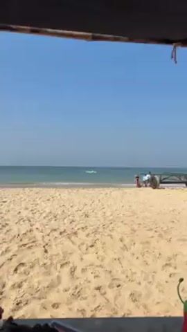 The scene is a sunny beach with a cloudless blue sky. In the foreground is a sandy beach. A woman in a black bikini is standing on the beach, facing the ocean. In the distance, a person is riding a jet ski in the ocean. To the right of the woman, a person is sitting on a tractor. The video appears to be a static shot.