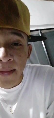 The video shows a young man wearing a yellow baseball cap and a white t-shirt. He is standing on a balcony at night, with city lights visible in the background. He points a finger towards the camera. The man is wearing a necklace with a cross pendant.