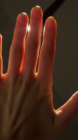 A pale hand is held up against a light source. The light shines between the fingers and around the edges of the hand, creating a bright outline. The background is dark.