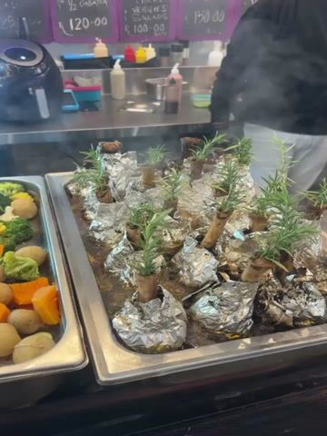 The video shows two metal trays containing prepared food. The tray on the left contains a medley of vegetables including broccoli, cauliflower, carrots, and potatoes. The tray on the right appears to hold individual servings of meat wrapped in foil, with a sprig of rosemary placed on top of each. Steam is rising from the food, indicating that it is freshly prepared or kept warm. In the background, a stainless-steel counter is visible with an air fryer and assorted condiments or ingredients. A menu board hangs above the counter displaying prices. A person is partially visible on the right side of the frame. The scene appears to be in a food service setting, possibly a buffet or cafeteria.