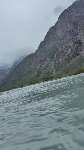 The video captures a wide shot of a river flowing through a mountainous landscape under an overcast sky. The river dominates the foreground, with its surface showing ripples and movement. The background features a steep, rocky mountain slope on the right, sparsely covered with green vegetation. Further back, more mountains recede into the distance, partially obscured by thick, low-hanging clouds. The overall atmosphere is moody and somewhat dramatic due to the cloudy weather and the imposing mountain terrain.