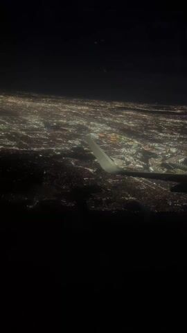 The video shows a nighttime view of a city from an airplane window. The city is illuminated with many lights, creating a sparkly, expansive scene. Part of the plane's wing is visible in the foreground. The sky above the city is completely dark.