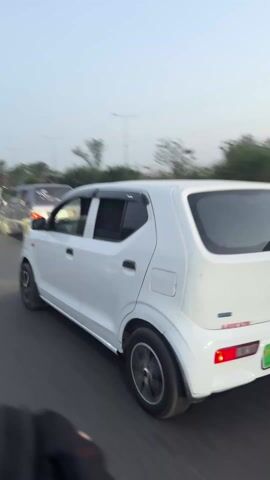 The video shows a white Suzuki Alto driving on a road in Pakistan. The car is traveling in the right lane and is followed by a blue motorized rickshaw. The sky is overcast, and there are trees and light poles lining the road. The camera is mounted on a vehicle traveling in the opposite direction.