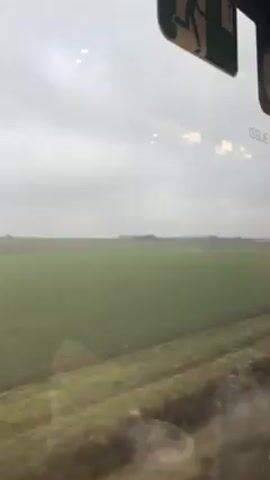 The video shows a view from a train window. Outside, there is a flat, green field stretching to the horizon under an overcast sky. An emergency exit sign is visible inside the train, partially obscuring the view. The window is slightly dirty with condensation or droplets. The camera is still and does not move.