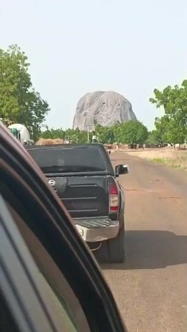 The video, shot from within a car, shows a black Nissan pickup truck driving on a paved road. The road has some patches of rough surface. In the background, a large, grey, rounded rock formation dominates the landscape. There are scattered trees and low-lying vegetation along the road. As the video continues, the view shifts, focusing more on the rock formation in the distance. The sky is a pale blue.