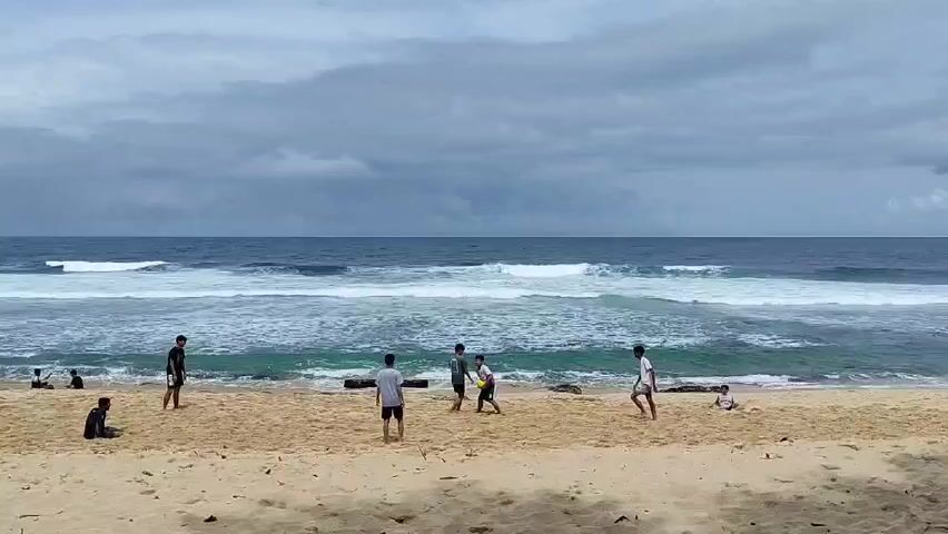 Here is a description of the video:

A group of young people are playing what appears to be soccer or football on a beach. They are kicking a red ball around on the sand near the shoreline. Some are running and making plays, while others are standing around watching. There are some other individuals scattered along the beach. Some appear to be relaxing or watching the game. The scene is set on a sandy beach with moderate waves rolling in from the ocean. The sky is overcast with dark clouds, suggesting potentially inclement weather. There is some driftwood on the sand.