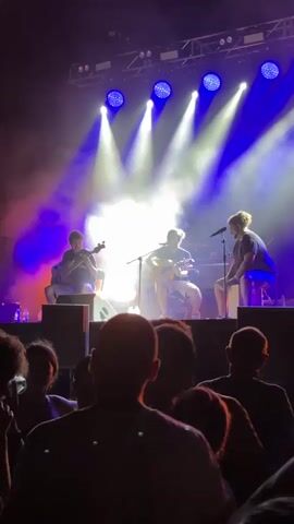 The video shows a live music performance on a stage. Three musicians are visible: one sitting on the left playing an instrument that appears to be a cello or upright bass, another in the center playing an acoustic guitar, and a third on the right, seated, with a microphone in front of them, possibly singing or speaking. The stage is lit with bright spotlights, predominantly blue and white, with some purple and pink hues. Smoke or haze is present on stage, catching the light. In the foreground, the backs of a crowd of spectators are visible, looking towards the stage. The overall environment is that of an indoor or outdoor concert venue.
