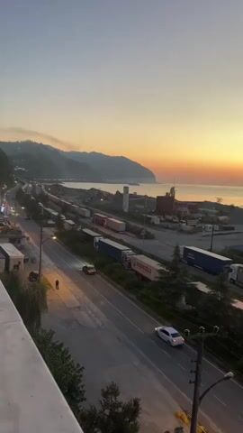 The video captures a scenic view of a coastal highway at sunset. The sky is painted with warm hues of orange and yellow as the sun dips below the horizon, casting a soft glow over the landscape. In the foreground, a road stretches out, with a few cars and a lone pedestrian visible. To the right, a line of trucks and semi-trailers are parked, suggesting a busy transportation hub. Beyond the road, a body of water, likely the sea, meets the shore, with a distant coastline dotted with hills and industrial structures. The overall atmosphere is tranquil, with the natural beauty of the sunset juxtaposed against the human activity of the highway and industrial area.