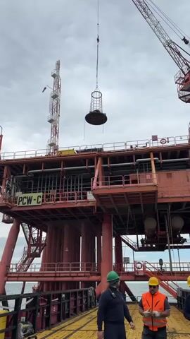 The image captures an offshore industrial scene under an overcast sky, likely near Marina di Ravenna, Italy. In the foreground, three men in work attire – hard hats and safety vests – are standing on a yellow-painted deck, observing an ongoing operation. A large, red and white crane, attached to an expansive, multi-level offshore platform, is actively lifting a large, cylindrical, cage-like object into the air. The platform itself is a prominent feature, with robust red/orange support legs and multiple decks. The text "PCW-C" is visible on one of the platform's structures. In the background, to the left of the main platform, another tall, slender metal structure, possibly a flare stack or communication tower, stands. The sea is visible beneath and around the platform, indicating its offshore location. The men appear focused on the crane's activity, suggesting a supervised lifting operation.