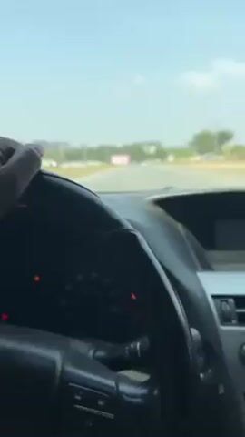 The video shows a view from inside a car, looking out through the windshield. The driver's left hand is visible on the steering wheel. The dashboard is visible with various controls and gauges. The car appears to be driving on a road with light traffic, buildings and trees are visible in the background under a clear, bright sky. There is a pink billboard visible in the distance. The general lighting suggests it is daytime.