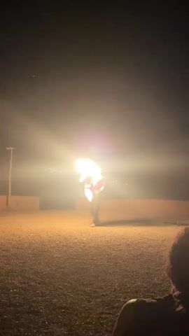 A person is performing with fire at night. They are standing in a dirt area with some low concrete walls behind them. The person is holding a torch in their right hand and appears to be breathing fire, with a large flame coming from their mouth. There are also flames on the torch. In the background, there are lights from a distant town or city. The sky is dark.