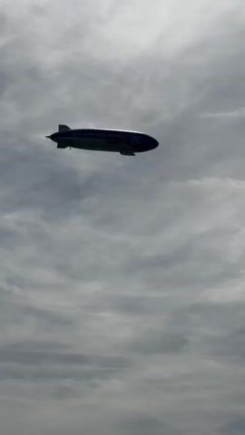 The video shows a blimp flying in the sky. The blimp is dark in color and appears to have some writing on its side. The sky is cloudy with some patches of sunlight. In the lower part of the video, there is a pier or dock with people, bicycles, and a sign that reads "Fahrkarten" which means tickets in German. There are also boats docked at the pier. The overall scene suggests an outdoor location, possibly a tourist area, on a day with mixed weather conditions.