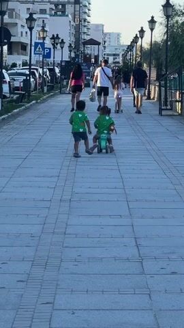 This media file shows a scene on a pedestrian walkway in Năvodari, Romania. The time of day appears to be late afternoon or early evening, with the sun casting long shadows. The weather seems pleasant, with clear skies.

The main subjects are several people walking along the path. In the foreground, two young children are engaged in activity. One child is walking, while another is riding a small, green toy vehicle, possibly a scooter or a tricycle. Both children are wearing green t-shirts with a clover leaf pattern. They are walking away from the camera, towards a group of adults.

Further down the path, several adults are walking. One adult, a woman in a pink top and dark shorts, is walking with the children. Behind her, a man in a white t-shirt and dark shorts, a woman in a dark top and shorts, and a man in a black t-shirt and shorts are also walking. Another young girl, in a pink top and white shorts, is on a scooter further ahead.

The setting is a paved walkway lined with lampposts and bordering a street with parked cars on one side and a low fence and vegetation on the other. In the background, there are modern buildings, suggesting an urban or resort area. Visible text includes a "P" sign for parking and a pedestrian crossing sign. The overall atmosphere appears relaxed and casual, with families enjoying an evening stroll.