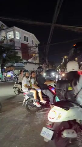The video captures a nighttime scene on a busy street, likely in Southeast Asia, judging by the vehicles and the tropical atmosphere. The foreground is dominated by a light-colored scooter, with a person wearing a white helmet and a light-colored top riding it. The scooter has a license plate visible. Behind this scooter, another scooter carries three passengers: a person in a helmet and two younger individuals. They are all looking in different directions. In the background, a large white bus with pink and red accents is visible, its lights illuminating the street. Further back, a white car and a silver car are also present. The environment appears to be urban, with buildings, overhead wires, and streetlights visible. The overall impression is one of traffic, movement, and nighttime activity.