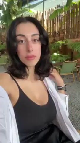 A woman films herself with a handheld camera. She has long, dark, wavy hair and dark eyes. She is wearing a black tank top and a white button-up shirt. She is wearing a silver bracelet on her left wrist and a smartwatch on her right wrist. The background shows a covered patio with plants, trees, string lights, and outdoor chairs. The camera moves slightly as she runs her fingers through her hair and adjusts the front of her shirt.