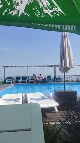 The video pans across a poolside scene on a sunny day. In the foreground, a white lounge chair is partially visible. Behind it, a swimming pool with clear blue water is lined with lounge chairs, some occupied by people. A large green umbrella with white text is visible in the upper portion of the frame, along with a closed white umbrella and an open white umbrella. People are scattered around the pool area, some sitting on lounge chairs and others in the water. The background shows a railing and a distant horizon with a hazy sky. Lavender plants are visible in the lower right corner of the frame.
