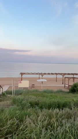 The video presents a serene beach scene under a pastel sky. The calm sea stretches horizontally to the horizon, meeting the sky in a soft gradient of light blues and pinks. In the foreground, green vegetation is visible, blurring slightly as the camera focuses on the sandy beach. The beach is sparsely populated with wooden structures, including a pergola-like frame and a sun umbrella. A small changing booth and a walkway made of wooden planks are also present. The overall scene is peaceful and quiet, with no visible people or activity.